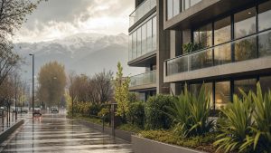 Edificio residencial moderno de hormigón con balcones de vidrio, fotografiado desde la calle en un día lluvioso, con cordillera nevada de fondo y vegetación en primer plano.